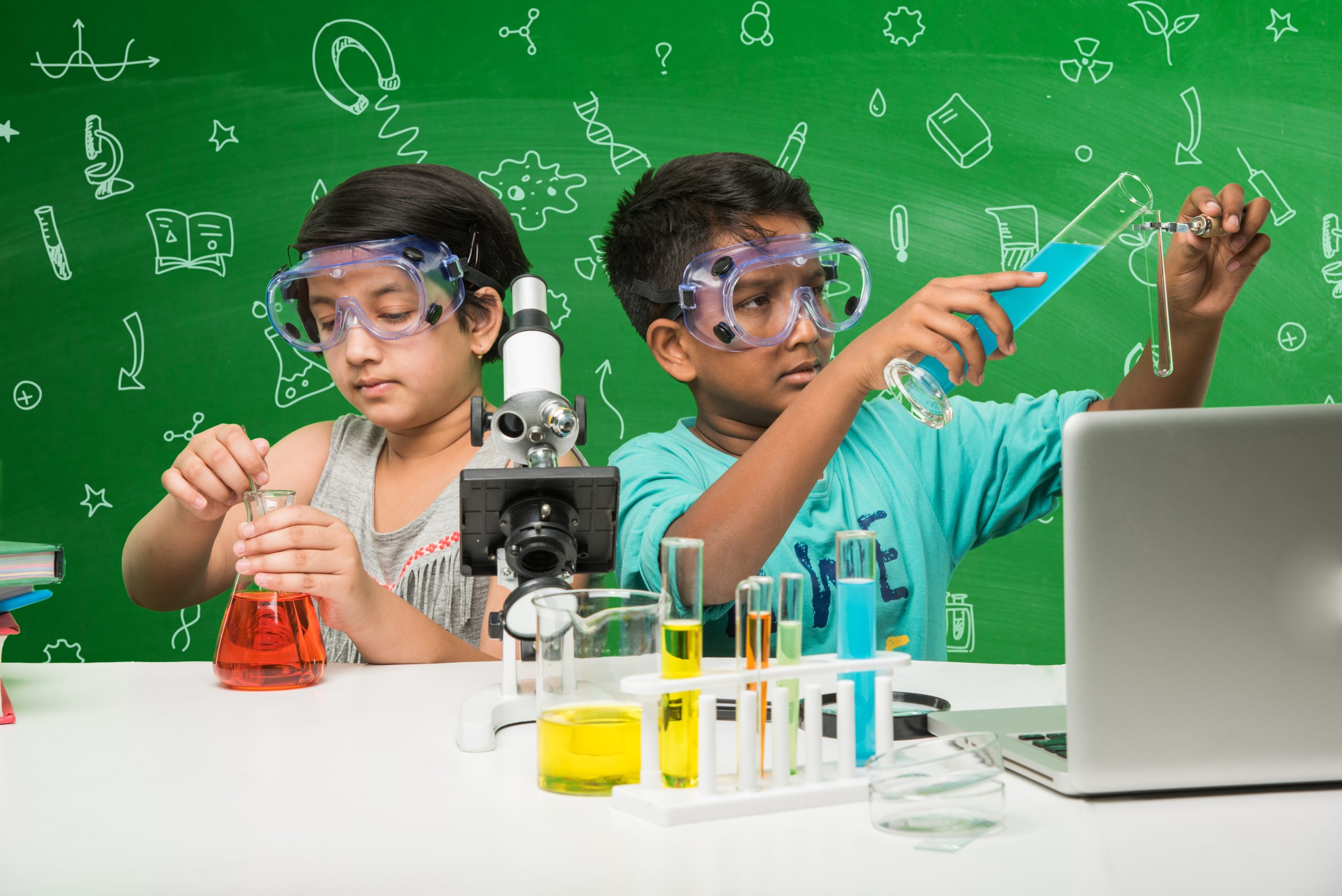 Kids and science concept - cute indian little kid student or scientist studying science or experimenting with microscope and Chemicals with diagrams doodles drawn over green chalkboard